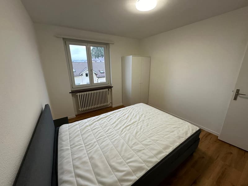 Spacious bedroom with white walls, hardwood flooring, a large window, a double bed, and a wardrobe cabinet.
