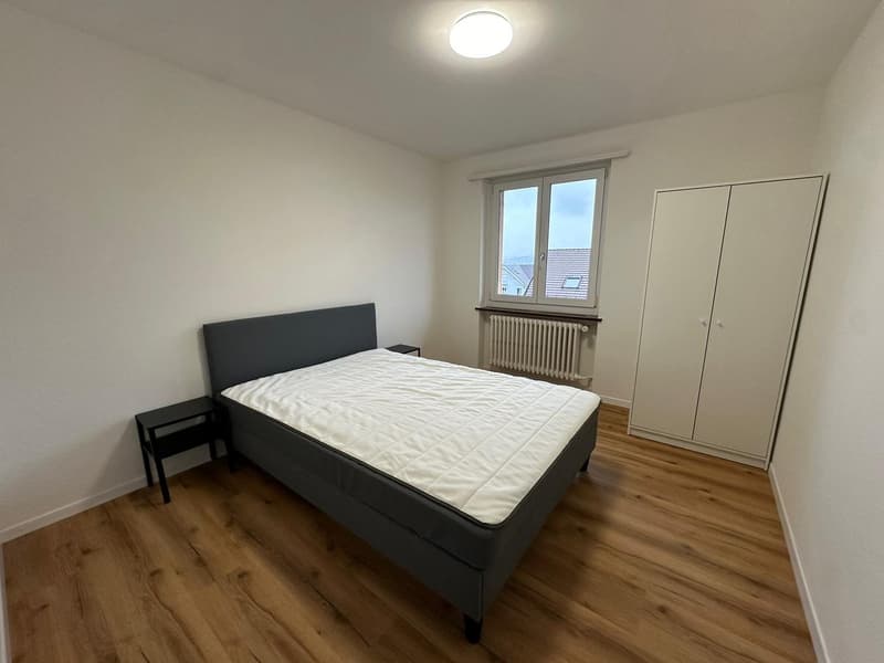 Spacious bedroom with hardwood flooring, white walls, a large window, a black bed frame with a white mattress, and a wardrobe cabinet.