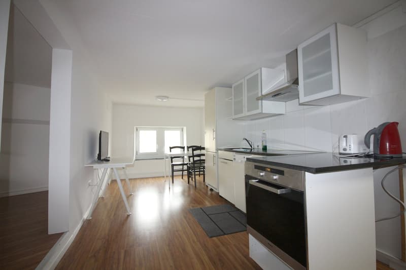 Open-concept kitchen and living room with hardwood floors, white walls, modern kitchen appliances including a dishwasher, dining table, and living room furniture