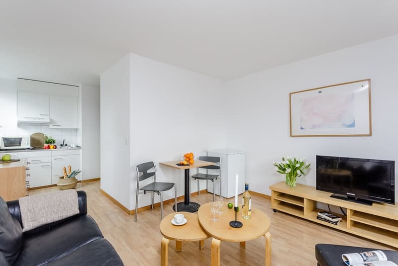 Minimalist living room with wooden floors, black leather couch, a round table, two chairs, TV, wine bottle, and a small candle