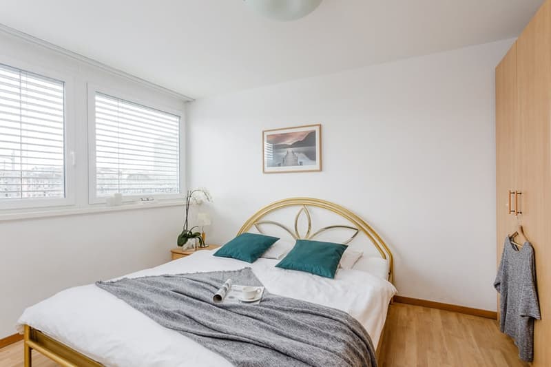 Spacious bedroom with large windows, white walls, wooden floor, gold-colored metal bed frame, bedside table, and a framed artwork on the wall.