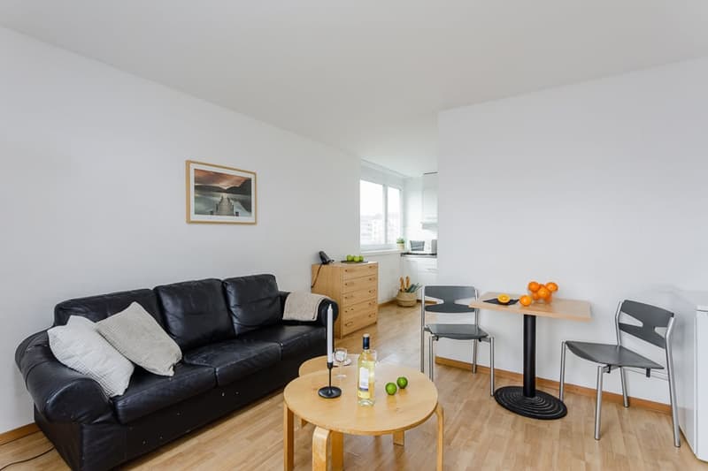 Spacious living room with hardwood floors, white walls, large windows, black leather sofa, round wooden coffee table, and a small dining area with a table and chairs