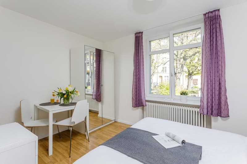 single bed, desk, chairs, radiator, mirror, window with view outside, table with flowers