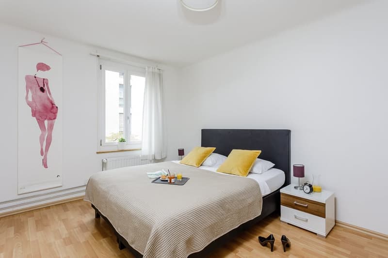 Spacious bedroom with hardwood flooring, white walls, large windows, black upholstered bed frame, bedside tables, and a decorative wall art piece.