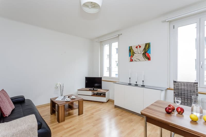 Bright and spacious living room with hardwood floors, white walls, large windows, modern furniture including a sofa, coffee table, and TV stand, and a colorful abstract painting on the wall.