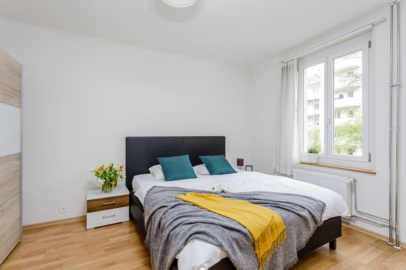 Spacious bedroom with white walls, hardwood flooring, large windows, a black upholstered bed frame, bedside tables, and a yellow throw blanket on the bed.