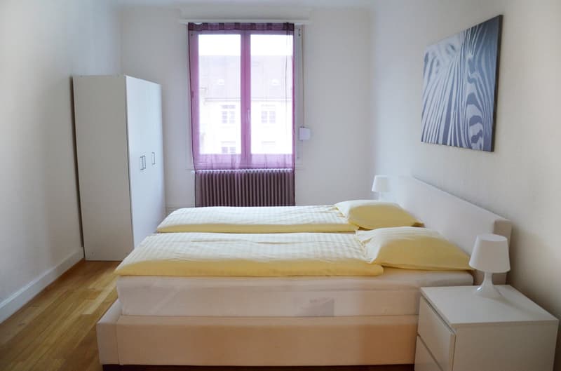 Spacious bedroom with white walls, hardwood floor, large window with purple curtains, white bed with yellow bedding, white wardrobe, and a painting on the wall.