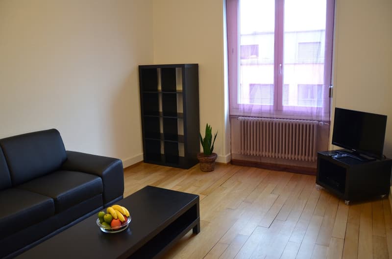 Spacious living room with hardwood floors, large windows, black sofa, coffee table, bookshelf, and a potted plant