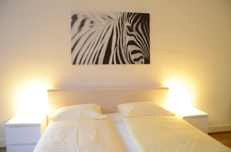 The image shows a bedroom with a white bed, white bedding, and a large black and white zebra-patterned artwork hanging on the wall above the bed. The room has a warm, cozy atmosphere with the soft lighting.