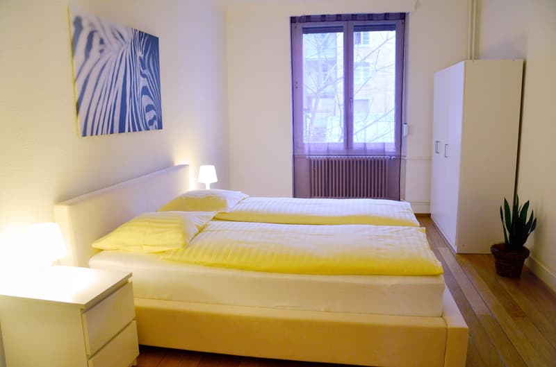 Bright and airy bedroom with white walls, large windows, a yellow bed with bedding, a nightstand with a lamp, and a wardrobe.