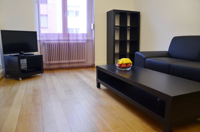 Spacious living room with hardwood floors, large windows, black TV stand, black bookshelf, and a blue sofa. A bowl of fruit is placed on the coffee table.