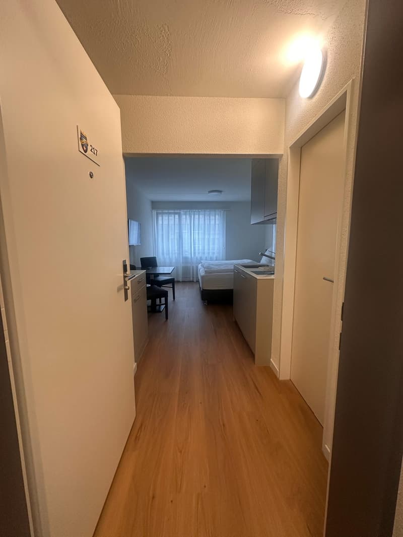 The image shows a studio apartment with a hallway leading to the main living area. The hallway has a numbered door and hardwood flooring. The main room appears to have a bed, desk, and kitchenette area.