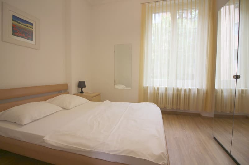 Spacious bedroom with a large window, wooden floor, and a framed artwork on the wall. The room has a double bed with white bedding, a bedside table, and a floor lamp.