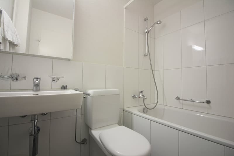 White tiled bathroom with a sink, toilet, and bathtub with a shower head. The bathroom appears clean and modern.