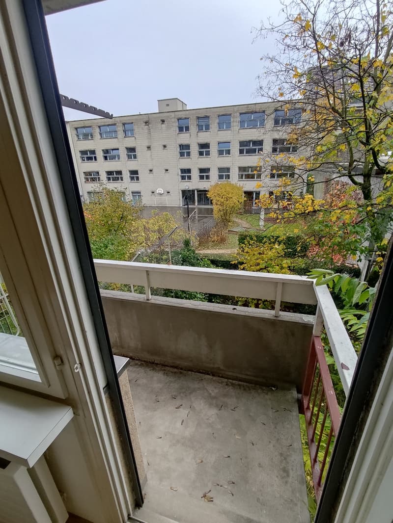 The image shows a multi-story building with a balcony or terrace visible from the window. The building has a concrete exterior and is surrounded by trees and greenery.