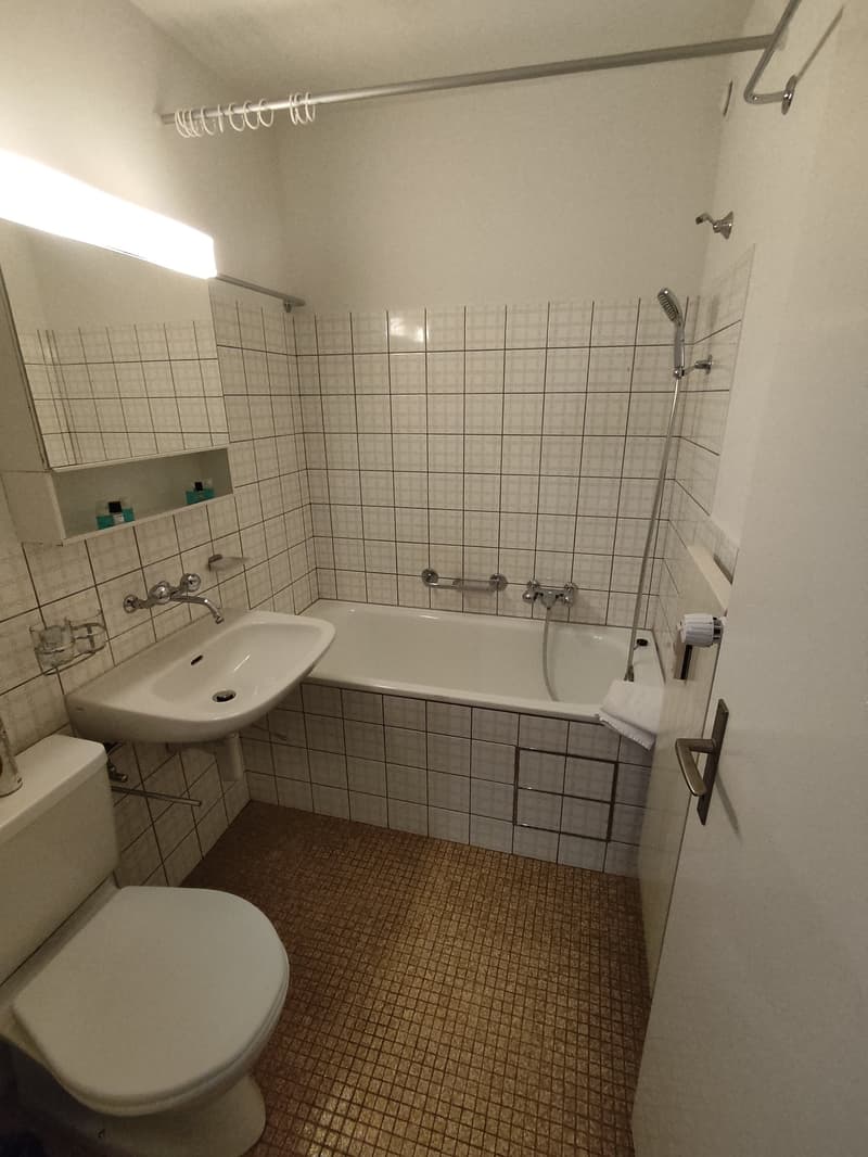 Tiled bathroom with a bathtub, sink, and toilet. The walls are covered in white tiles arranged in a grid pattern. There is a shower curtain rod above the bathtub.