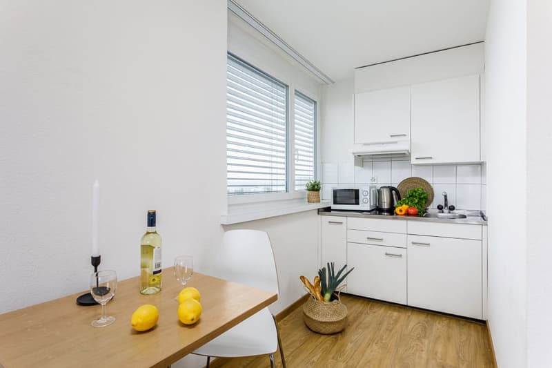 modern kitchen, white cabinets, wooden floor, dining table with lemons, wine bottle, white chair, large window, microwave, kettle, sink, potted plant, baskets with vegetables and bread
