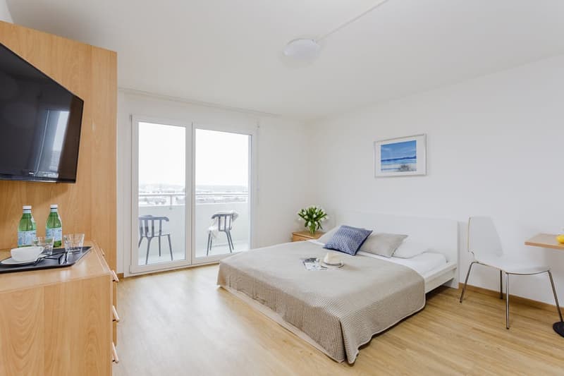Wooden floor, white walls, sliding glass doors to balcony, large bed, bedside table, flowers, chair, artwork, television