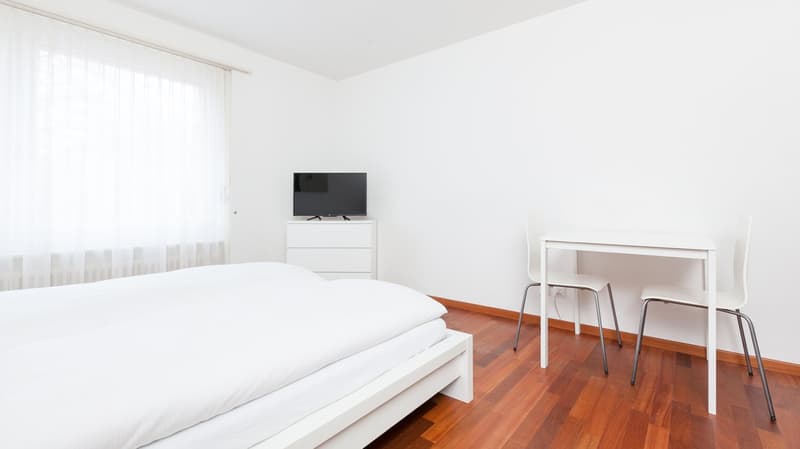 Bedroom with white walls, bed, TV stand, two chairs, wooden floor, white bed, white desk, white chair, white curtains, white sheet.