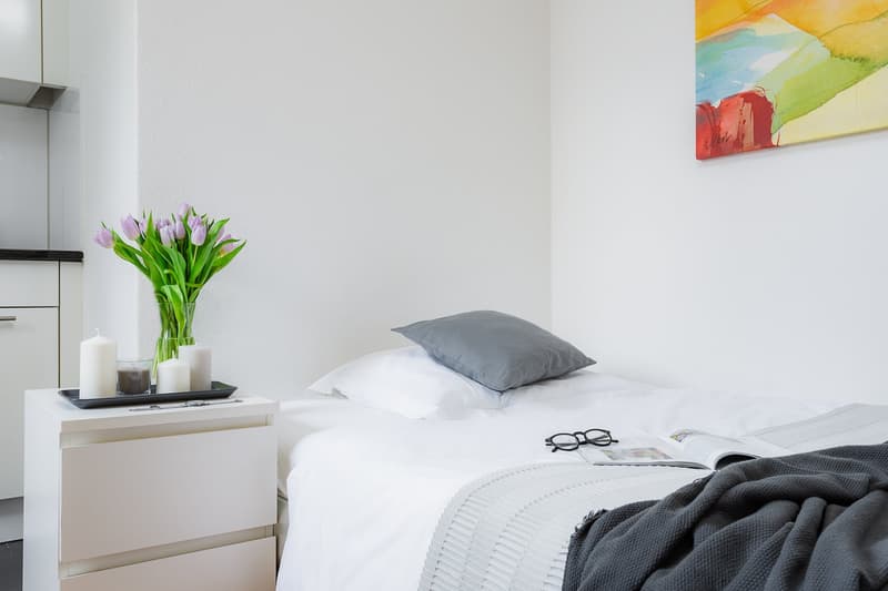 Modern minimalist bedroom with white walls, white furniture, a single bed, nightstand, and a painting on the wall.
