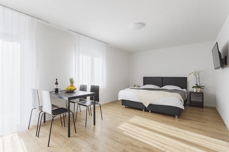 dining table with chairs, double bed with white blankets, white walls, television, floor lamp, flower vase, light wood floor