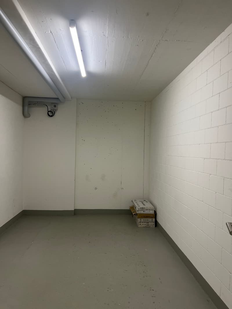 Empty room with white walls, white ceiling, grey floor, single ceiling light, and a few boxes in the corner