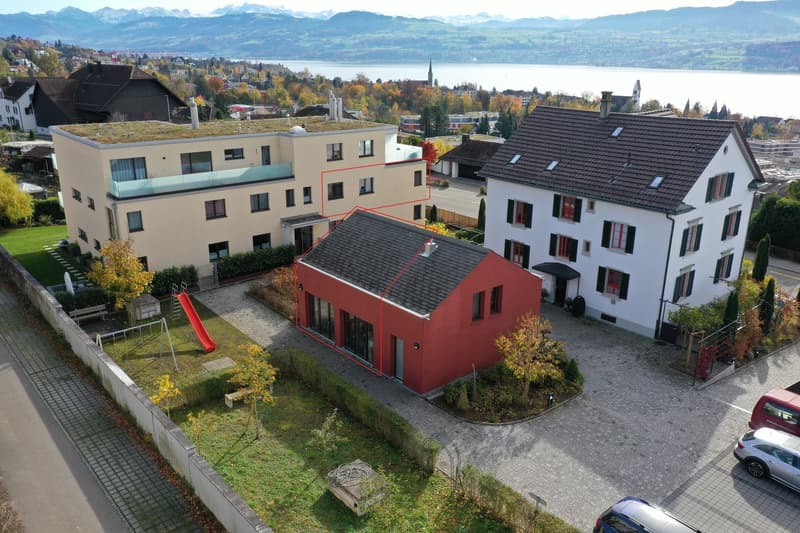 Modern single-family house, red exterior, large windows, highlighted in red, green rooftop, located in a neighborhood with other houses, lake and mountains in the background.
