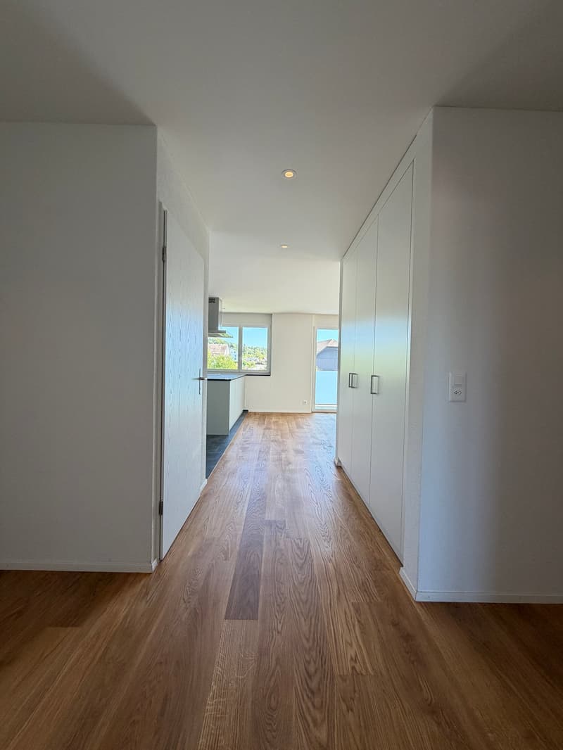 Empty hallway with wooden floor, white walls, large windows, and a kitchen area.