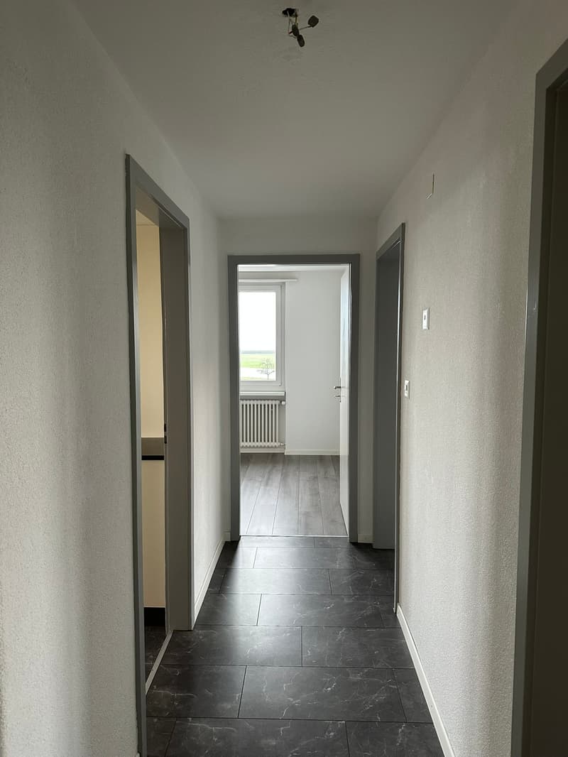 Empty hallway with white walls, black tiled floor, and gray doors. A window and radiator are visible in the room at the end of the hallway.