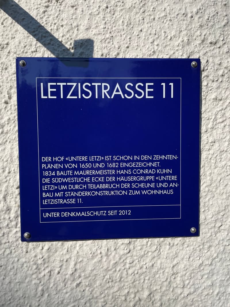 A blue informational plaque is affixed to a white wall. It contains German text that outlines the historical significance of a courtyard and a building located at Letzistrasse 11. The plaque mentions that the courtyard was first depicted in the 16th centu