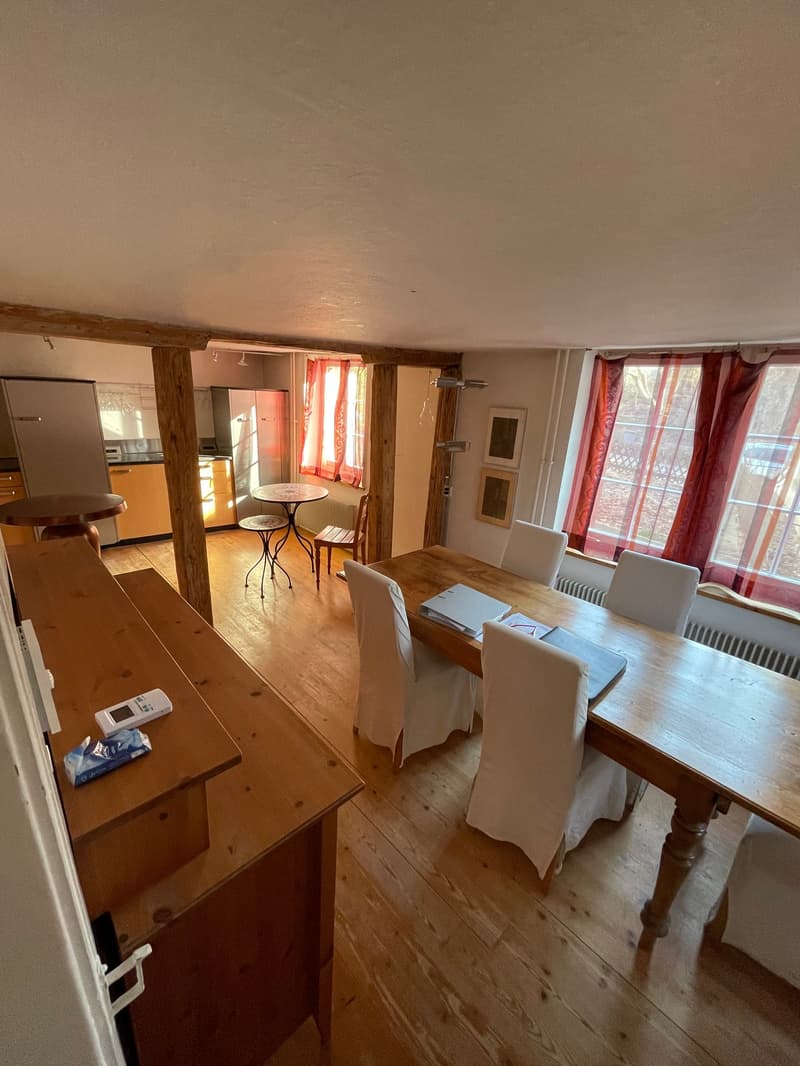 Dining area with wooden dining table, wooden cabinet, radiator, red curtains, two small tables and chairs, and refrigerator.