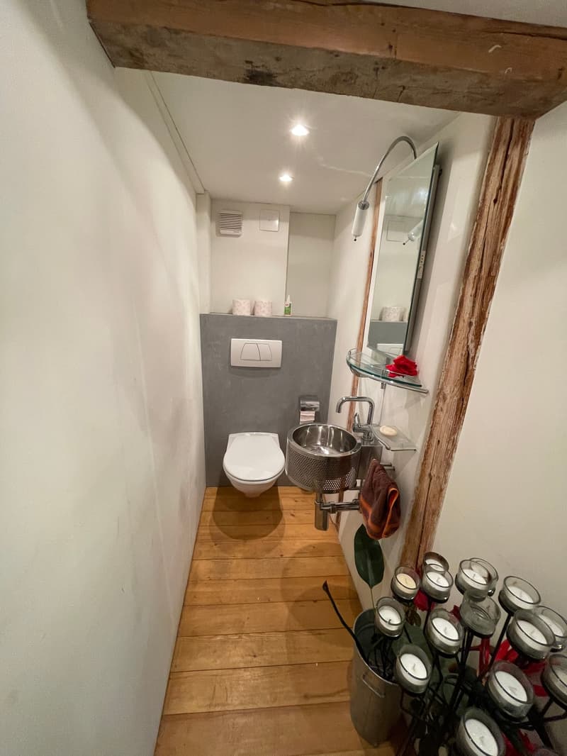 Modern bathroom with white toilet, grey tiled wall, silver sink with faucet, mirror, mounted towel rack, wooden floor, candles on holder