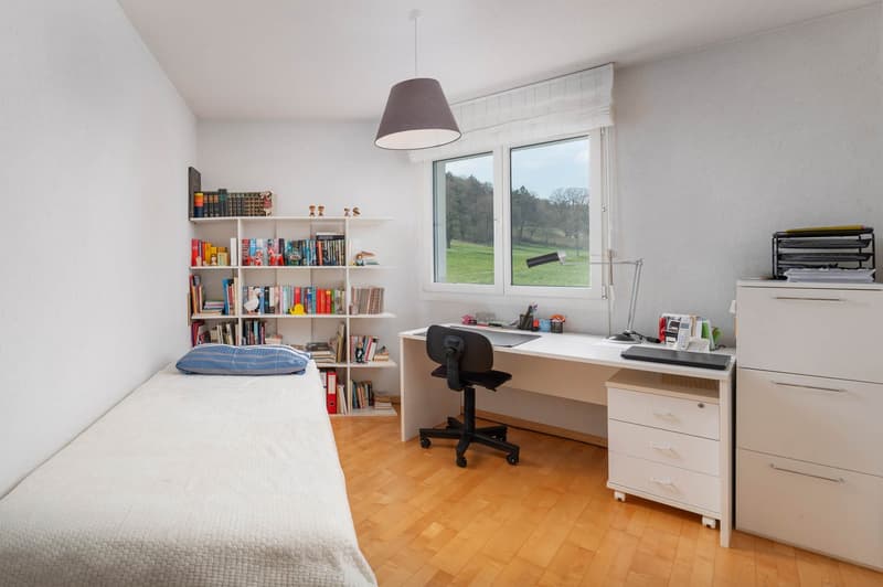 A small bedroom with a bed, a desk, a chair, a bookshelf, and a window with a view of a grassy field.