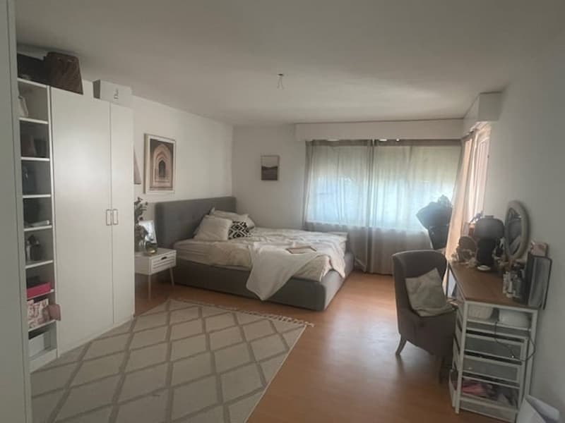 A bedroom with bed, bedside table, rug, chair, desk, picture frames, and a large window with curtains.