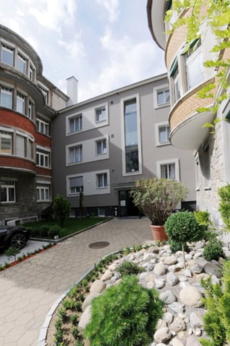 multi-story building with grey exterior, small garden, driveway, car parked