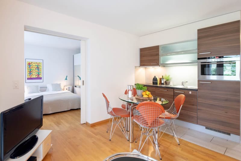 Modern open plan kitchen, dining area, living room and bedroom, hardwood floors, dining table with chairs, orange modern chairs, white television, bedroom with bed and pillows