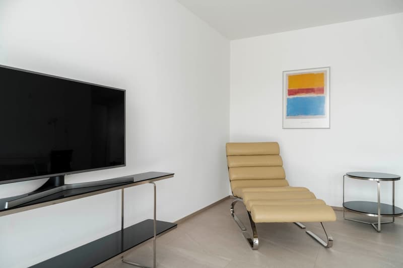 a modern living room with a black TV on a stand, a beige lounge chair, and a framed picture on the wall