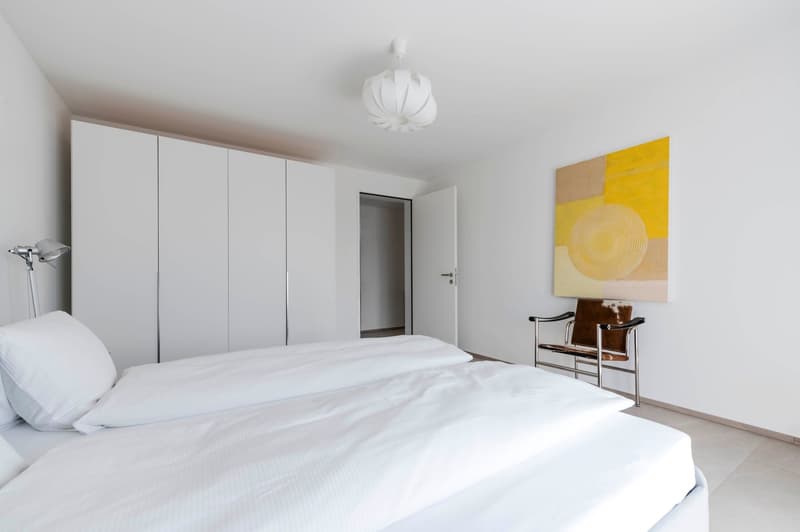 bedroom with two single beds, white walls, large white wardrobe, open door, lamp, painting, chair