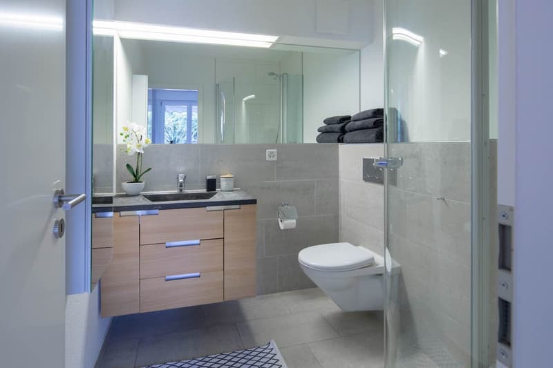 modern bathroom with white tiles, wooden cabinet, sink, toilet, shower enclosure, and mirror