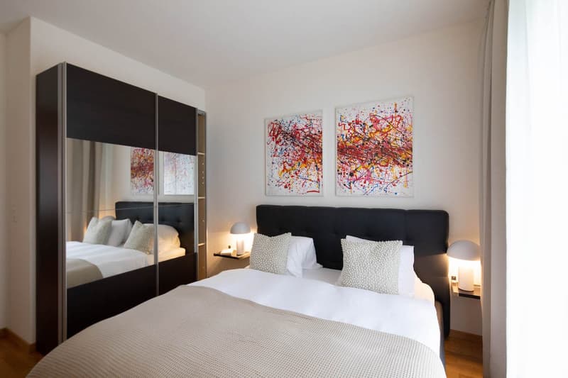 White walls, double bed with decorative pillows, two lamps, abstract paintings on the wall, mirror closet, wooden floor