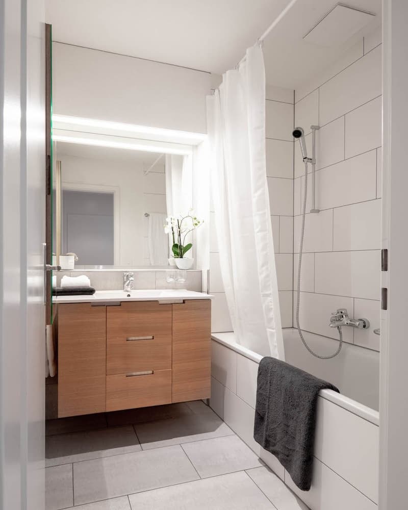 Modern bathroom with white tiles, wooden cabinet, white sink, mirror, flower vase, white shower curtain, towel, shower head, and bathtub