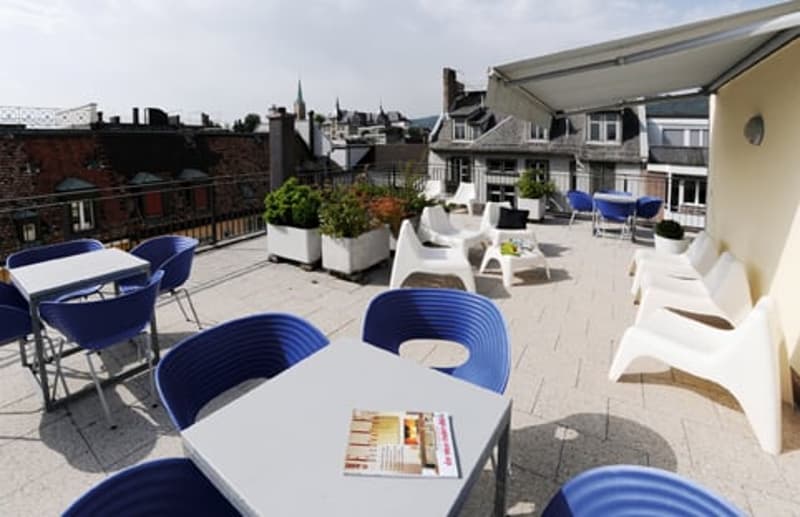 Outdoor seating area with blue and white chairs and tables, covered by a white awning, view of the cityscape, with plants and greenery.
