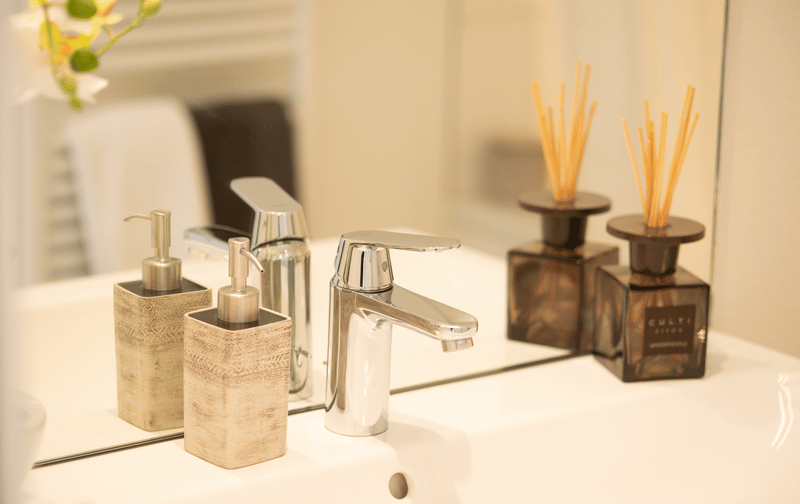 White counter top, silver sink, silver faucet, soap dispensers, and two bottles with incense sticks.