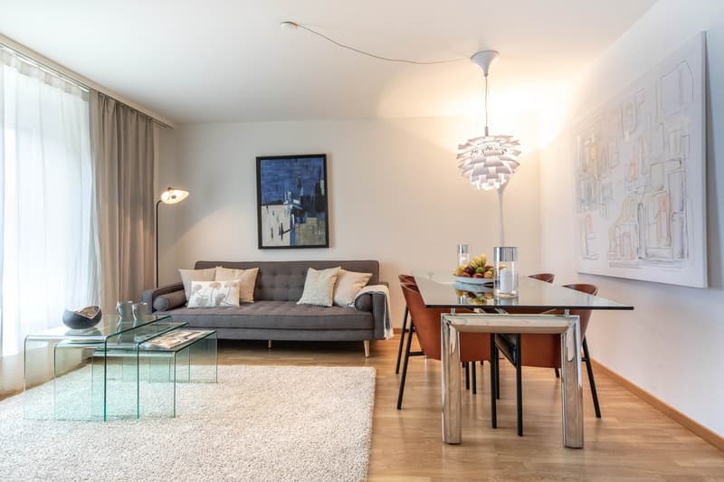 living room with a sofa, dining area with glass table and 4 chairs, rug, paintings, lamp, chandelier