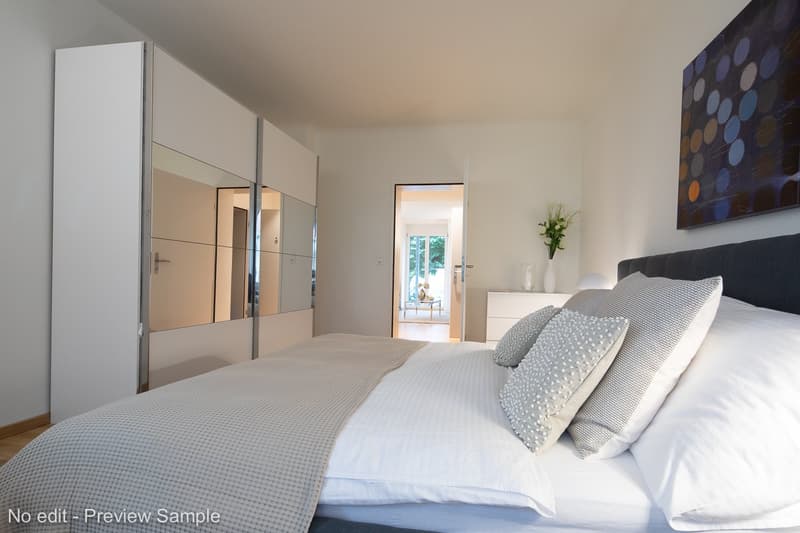 Modern bedroom with a double bed, wardrobe with mirror, nightstand with lamp, white linen, and a floral vase.