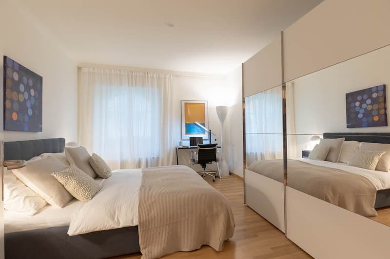 two beds, two nightstands, large window with curtains, paintings, desk, computer, chair, lamp, gray walls, hardwood floors