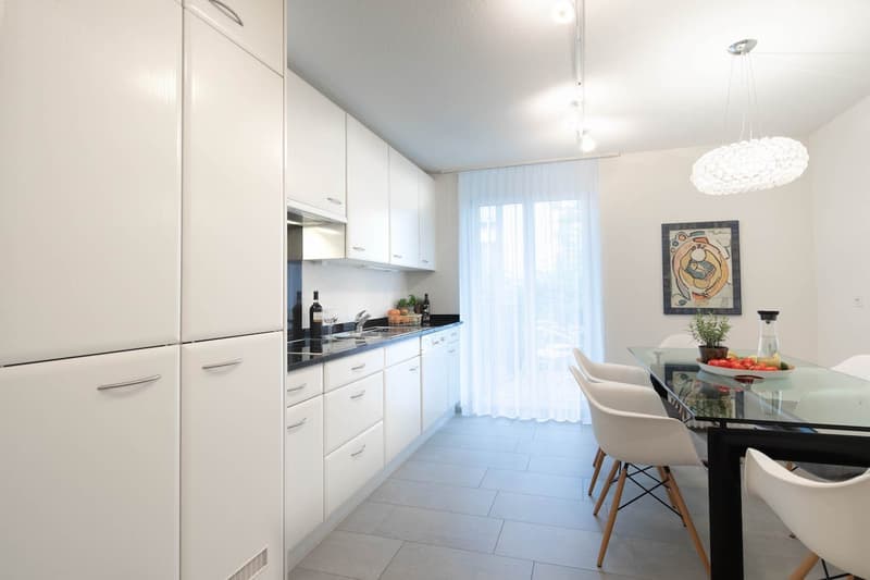 Modern kitchen with a dining area. White cabinets, black countertops, glass table, white chairs, a pendant lamp, a chandelier, a picture frame, and white curtains.