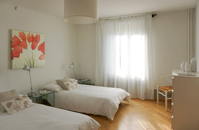 room has 2 double beds, lamps, nightstands, framed painting on wall, white curtains on window, wooden flooring