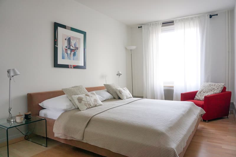 white walls, wooden floor, a bed with four pillows, a chair, night tables, lamps, a window with white curtains, a painting on the wall