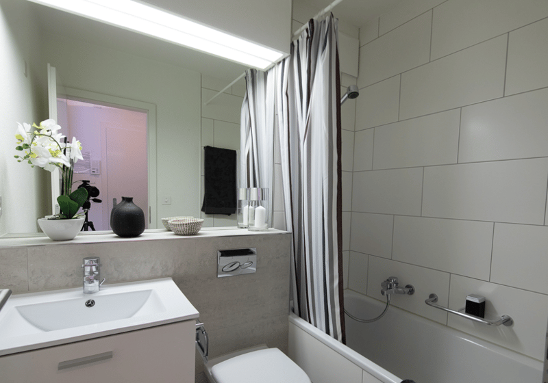modern bathroom with white tiles, white bathtub, white vanity, white sink, silver faucet, shower curtain, shelf with flowers and decor